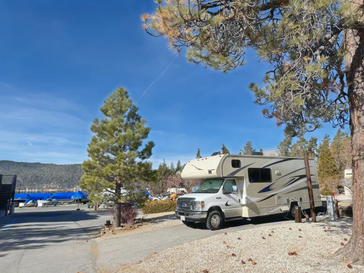 A white RV rental motorhome is parked at a sunny lakeside campsite, surrounded by pine trees with covered boats.. Winnebago Minnie Winnie 2020