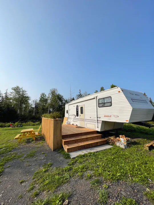 Your scenic rv rental awaits! A white camper with a wooden deck and picnic table, nestled among green trees.. Jayco Eagle 1998