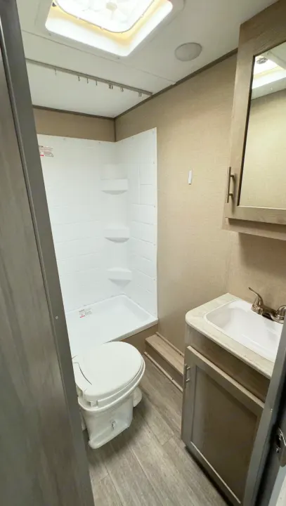 Clean RV rental bathroom with white shower, toilet, sink, vanity, mirror cabinet, and ceiling skylight.. Coachmen Catalina 2023