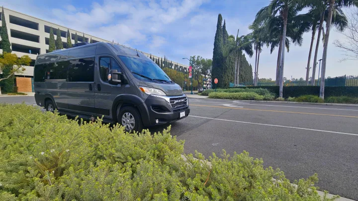 Grey Gazelle camper van, an RV rental, parked by green bushes. A large parking structure and tall trees are in the background.. Thor Motor Coach Dazzle 2025