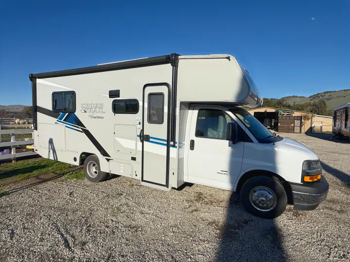A white Coachmen Cross Trail rv rental motorhome is parked on a sunny gravel lot, with green hills and a blue sky in the background.. 1HAGGUC78MN008793 Cross Trail 2022