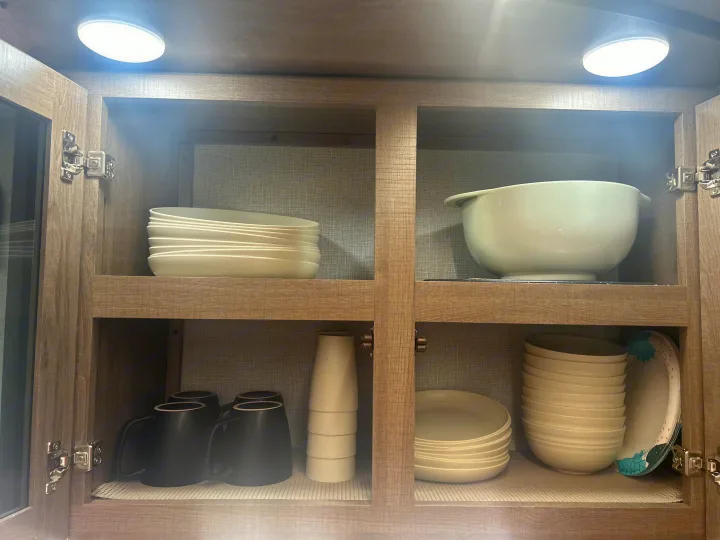 Inside an RV rental kitchen cabinet: stacked white dishes, mugs, and a mixing bowl on non-slip mats, illuminated by LED lights.. Grand Design Imagine XLS 2024
