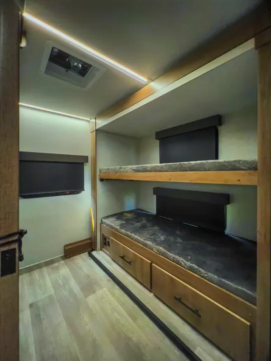 RV rental's bunk area: two beds with gray mattresses, storage drawers, wall-mounted TV, and bright LED lights.. Grand Design Imagine XLS 2024