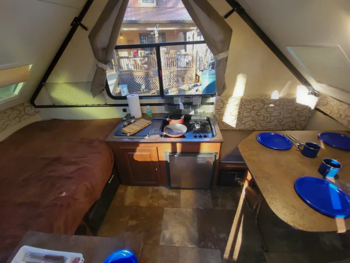 Interior of a compact RV rental, showing a cozy sleeping area, compact kitchen with stove and fridge, and dining table with blue dishes.. Flagstaff A Frame 2015