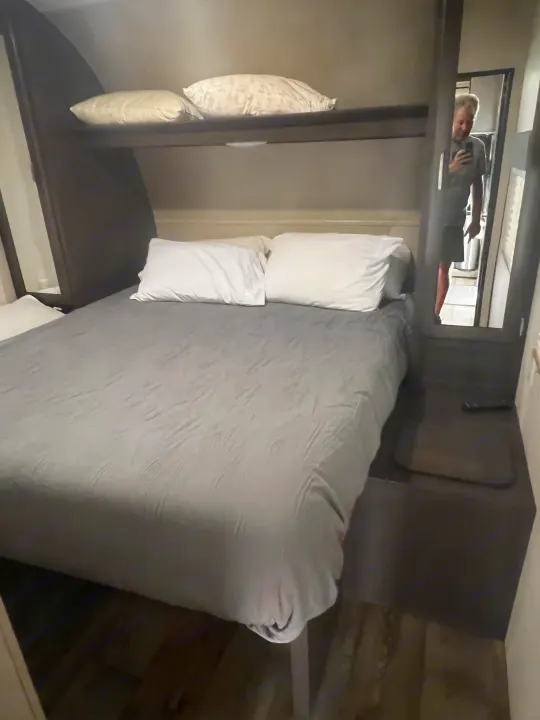 Cozy RV rental bedroom with a gray bed, white pillows, and overhead shelf. Mirror reflects person inside.. Forest River Sandstorm 2017