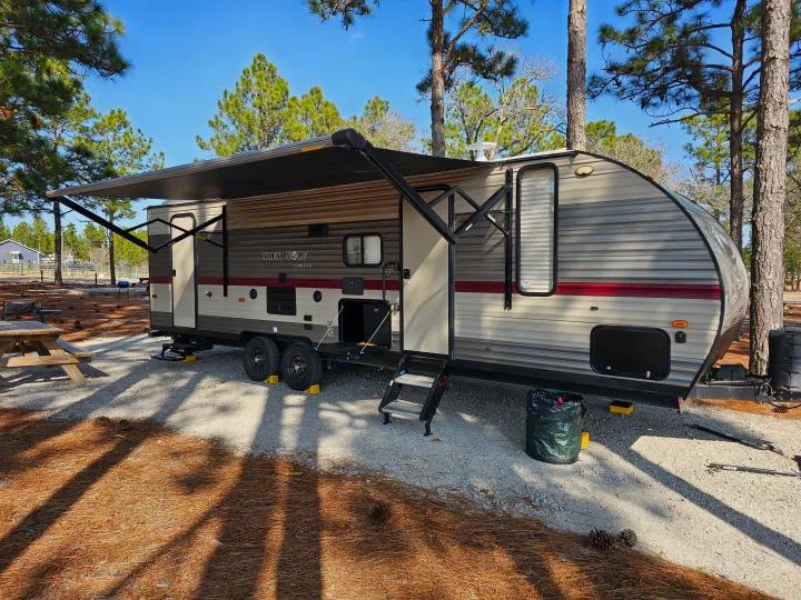 Your next RV rental destination: a cozy travel trailer parked in a serene pine forest campsite with an extended awning.. Forest River Cherokee Grey Wolf 2019