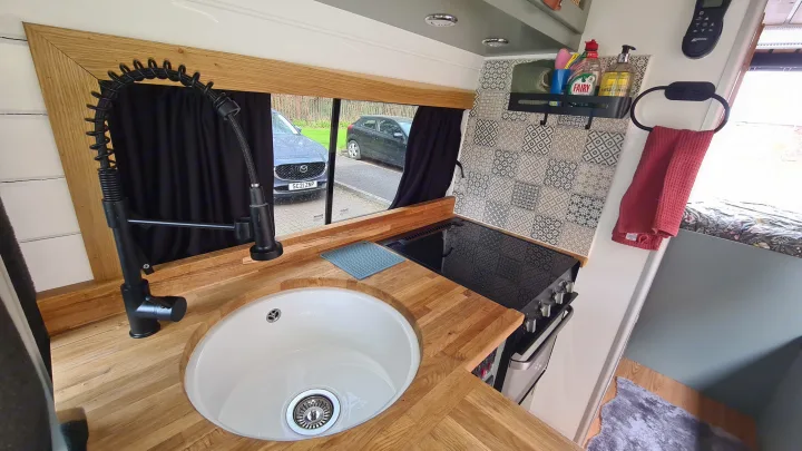 A sleek RV rental kitchen with a white sink, black faucet, patterned backsplash, wooden counters, and cooktop for travel.. Citroen Relay L3H2 2022