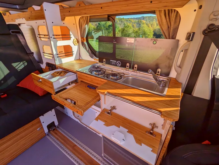 Inside a custom-built camper van rv rental, featuring a compact kitchen with a stove, sink, pull-out board, and comfy seating.. Renault Trafic 2025