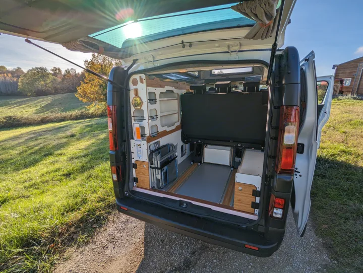 Inside a custom RV rental camper van with open tailgate, revealing smart storage, convertible bed, and chairs in nature.. Renault Trafic 2025