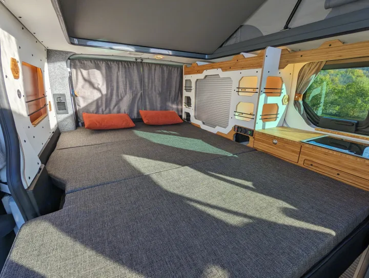 Rv rental camper van interior: a spacious grey bed with orange pillows, sleek wood kitchenette, and pop-top roof.. Renault Trafic 2025