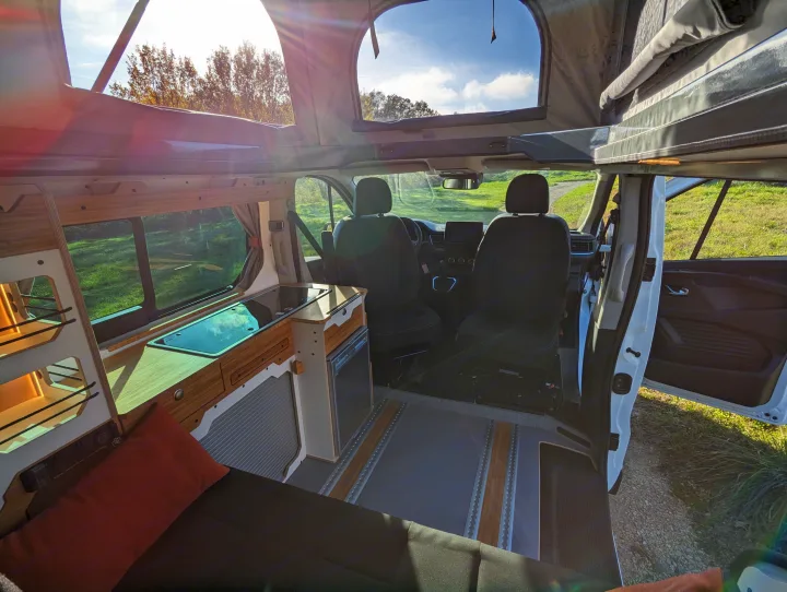 Bright campervan rental interior: pop-top open to sky, kitchenette, cozy bench, and front cabin view for road trips.. Renault Trafic 2025