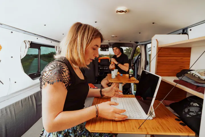 A woman uses a laptop on a bamboo table inside an RV rental. A man sits nearby. Perfect for remote work and van life adventures.. Renault Trafic 2025