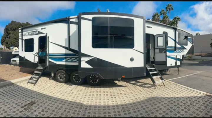 Spacious white and black fifth-wheel RV rental with open doors and steps, parked on pavers under a blue sky with palm trees.. Heartland RVs Road Warrior Toy Hauler 2022