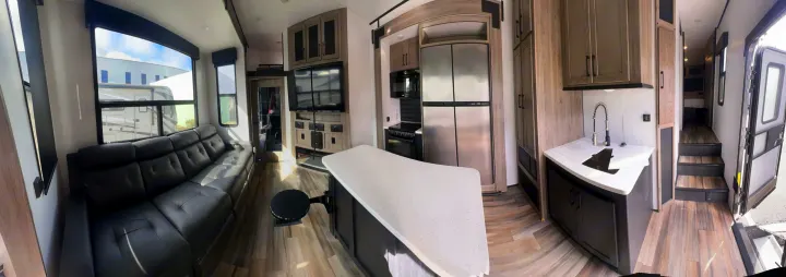 Panoramic view of a modern RV rental interior: spacious living room with leather sofa, island kitchen, and bedroom access.. Heartland RVs Road Warrior Toy Hauler 2022