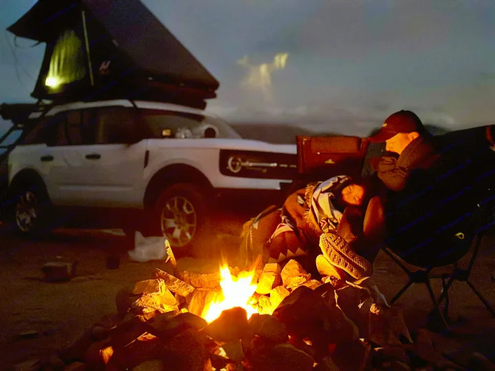 Enjoying an outdoor adventure with an **rv rental**-like setup. Campfire glows as two people relax by a white SUV with a rooftop tent.. Ford Bronco sport 2022