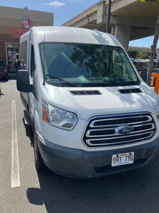 A white Ford Transit van, often used for RV rental, parked in Hawaii at Dunkin' under an overpass.. Ford Transit 2018