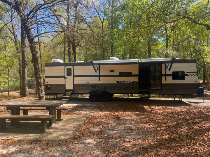 Spacious rv rental travel trailer set up in a sunny, wooded campsite with a concrete picnic table, ready for outdoor adventure.. Gulf Stream Innsbruck 2023