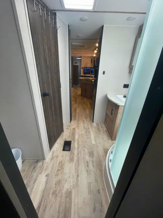 RV rental interior: View of a modern RV hallway with wood floors, revealing a bathroom with shower & vanity, a door, and a kitchen.. Gulf Stream Innsbruck 2023