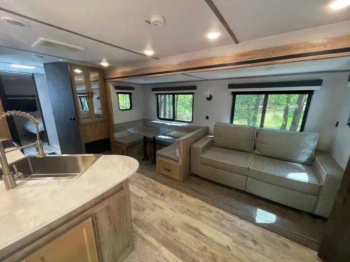 Modern RV rental interior showing a well-lit kitchen counter, U-shaped dinette with wood accents, and a comfortable grey sofa.. Gulf Stream Innsbruck 2023
