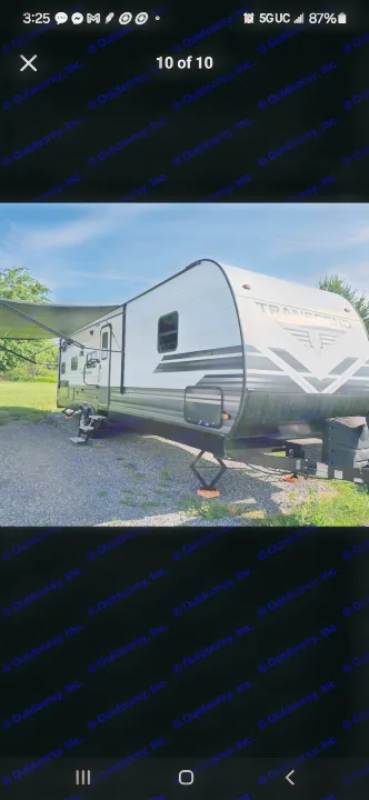 This clean white and grey travel trailer, ready for your next rv rental adventure, is parked on gravel with its awning extended.. Grand Design Transcend 2019