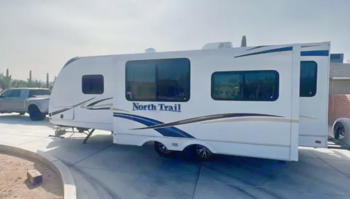 White North Trail travel trailer with extended slide-outs, parked on a driveway with a pickup truck nearby. Ideal for RV rental.. Heartland RVs North Trail 2011