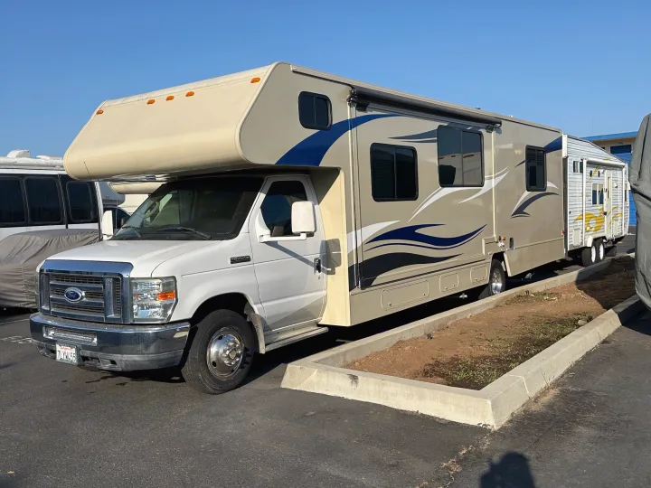 This Class C **rv rental**, a light tan motorhome with blue/white graphics, is based on a Ford E-series. It's parked outdoors.. Winnebago Minnie Winnie 2015