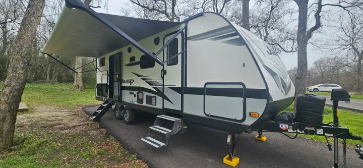 This white travel trailer RV rental, with an extended awning, is parked on an asphalt pad amidst trees and green grass.. Jayco Jay Feather 2022