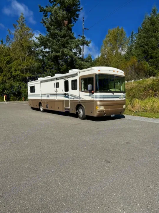 Spacious white & gold RV rental for adventure, parked on asphalt under a blue sky with trees.. Fleetwood 39/MH 2000