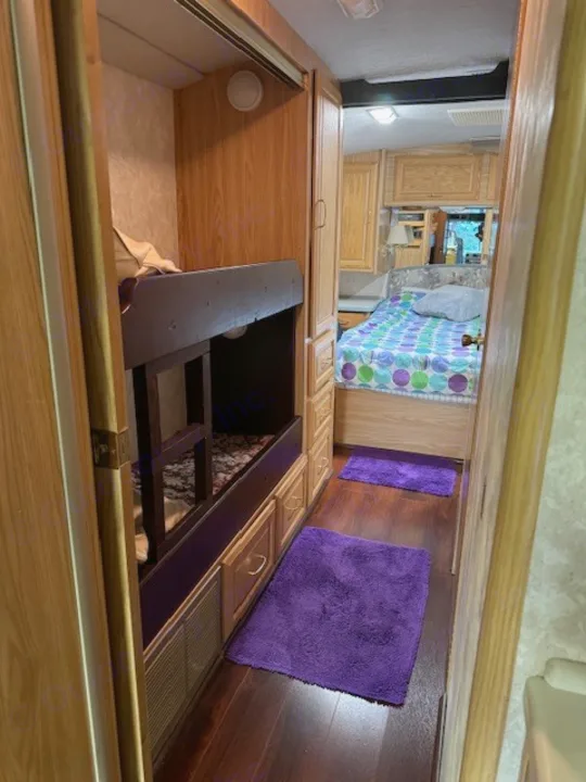 Cozy RV interior: bunk beds on left, wood-floored hallway with purple mats, main bedroom ahead. Ideal for an rv rental.. Fleetwood 39/MH 2000