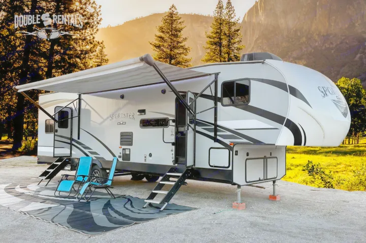 RV rental: A gray fifth-wheel camper with extended awning, outdoor mat, and chairs at a scenic mountain campsite, sunlit.. KZ Sportster 2021