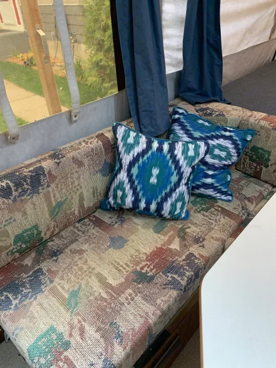 RV rental interior showing a patterned bench seat with blue and green pillows, blue curtains, and a white table.. Forest River Flagstaff 2000