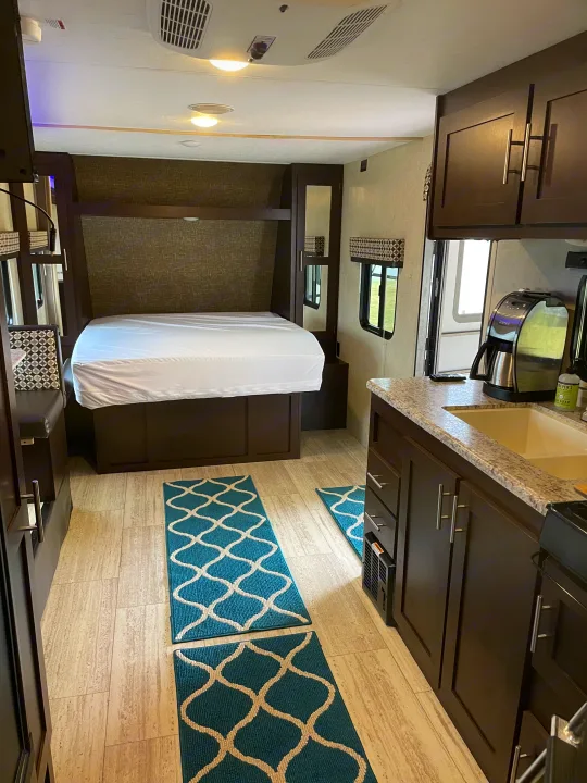 Experience a modern RV rental interior featuring a cozy bedroom, bright blue geometric rugs, and a compact kitchen setup.. Sportsmen Sportsmen Trailer 2017