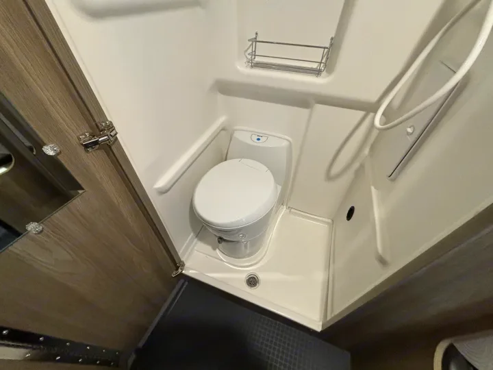 RV rental compact wet bath featuring a white toilet, shower, drain, and a small wall-mounted storage shelf.. Winnebago Solis NPF 2025