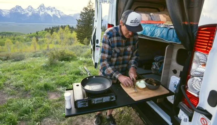 RV rental adventure: Man chops onions on an outdoor stove setup next to his camper van, stunning mountains beyond.. Winnebago Solis NPF 2025