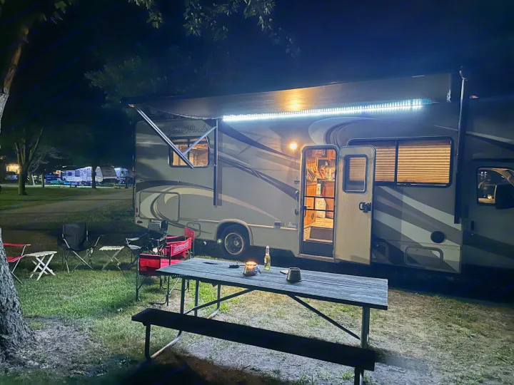 Brightly lit RV rental at a night campsite, featuring an illuminated awning, picnic table, and chairs for outdoor comfort.. Thor Four Winds 2015