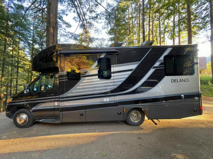 A sleek black and grey Delano motorhome, available as an rv rental, parked amidst sunlit forest trees.. Thor Delano Sprinter 2020