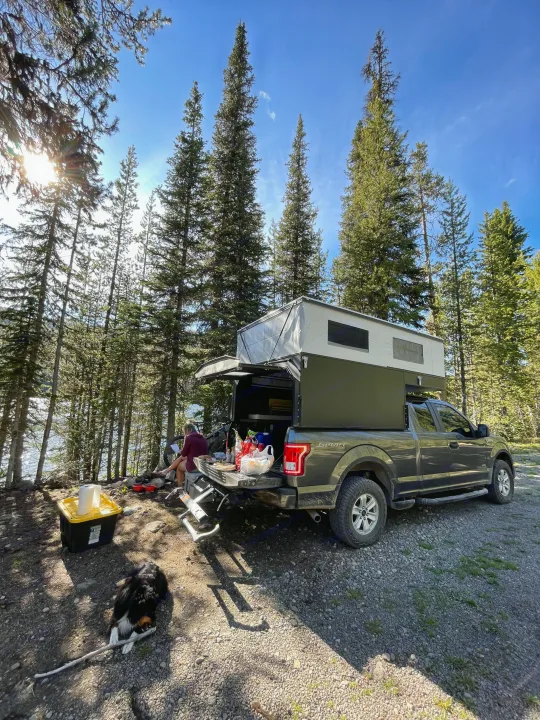 A person and dog relax by a lake next to a pop-top truck camper. Explore nature with an RV rental like this!. Four Wheel Campers Shell Models 2020