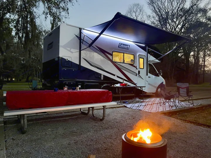 Relaxing outdoor evening with an rv rental at a cozy campsite, complete with a fire pit and illuminated awning.. Thor Coleman 2024