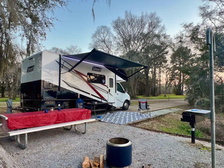 Spacious RV rental set up at a scenic campsite with an extended awning, picnic table, outdoor rug, fire pit, and grill.. Thor Coleman 2024
