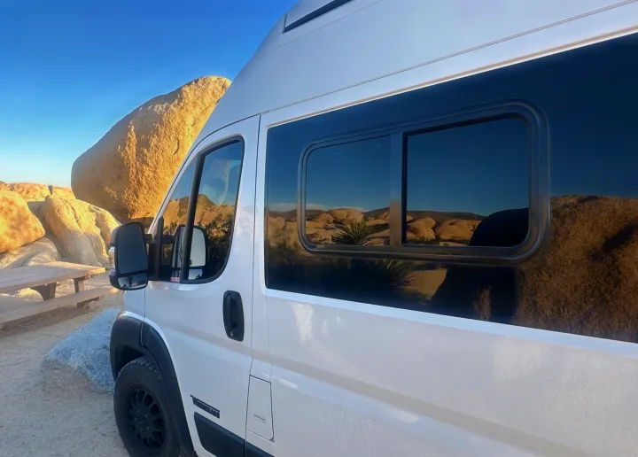 A white Winnebago camper van (RV) parked among massive, light-brown desert rocks, reflecting blue sky. Ideal for an rv rental.. Winnebago Solis 2023