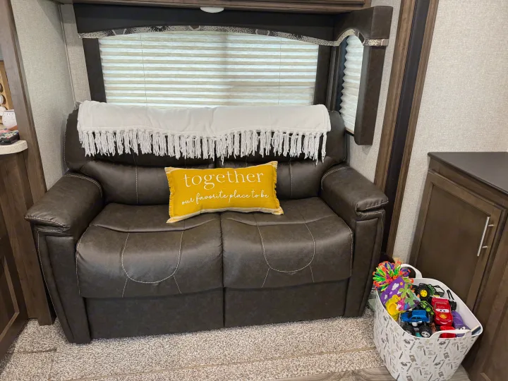 Comfy RV rental living space: a brown leather couch with a white throw, yellow pillow, and bin of toys.. Keystone RV Cougar X-Lite 2018