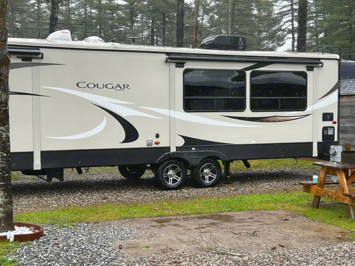 This Cougar travel trailer, perfect for an rv rental, is parked on a rainy gravel site, showing its beige exterior and slide-out.. Keystone RV Cougar X-Lite 2018