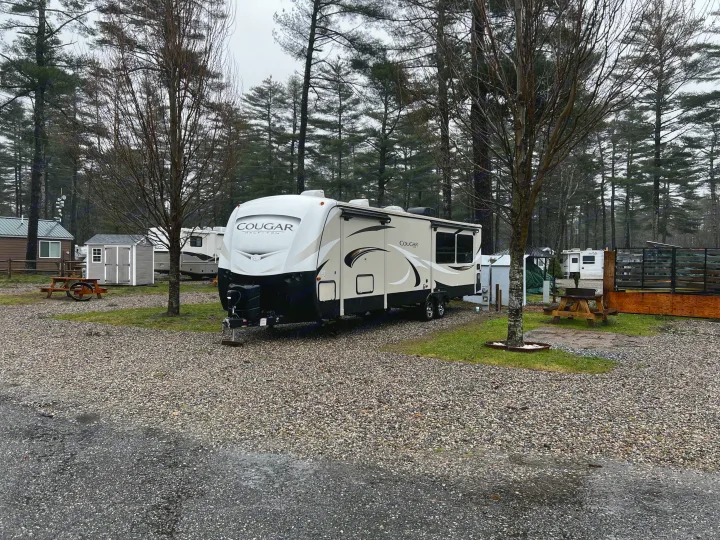 Experience an RV rental! A large travel trailer sits on a gravel lot at a shaded campground with cabins and picnic tables.. Keystone RV Cougar X-Lite 2018