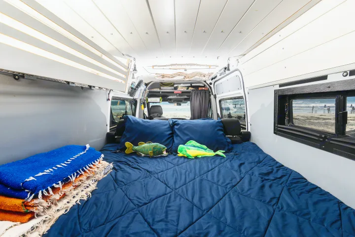 This cozy rv rental interior features a comfy bed with fish toys, stacked blankets, and open doors revealing a beach view.. Mercedes-Benz Sprinter 2018
