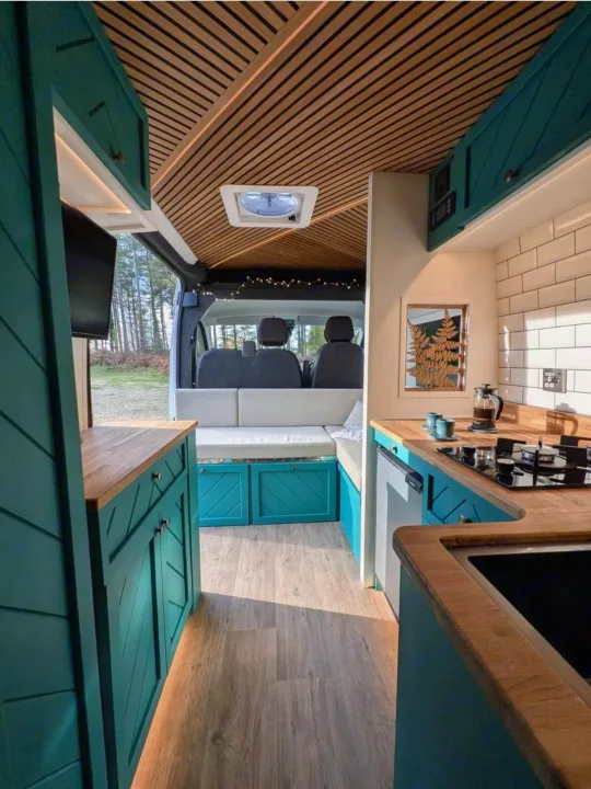 Inside a spacious rv rental camper van: teal custom cabinets, wood slats on ceiling, kitchen with stove/sink, comfy bench seating, forest view.. Peugeot Boxer L4H2 2021