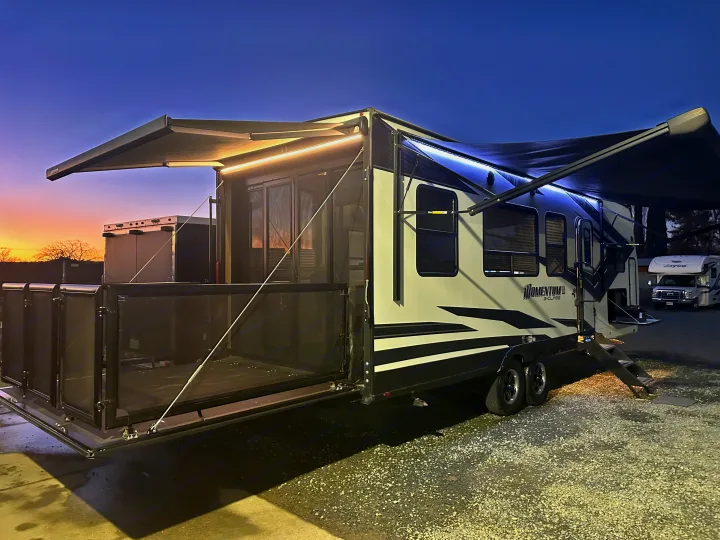 A sleek, black and white RV rental toy hauler, with illuminated awnings and a rear patio, set against a colorful dusk sky.. Grand Design Momentum 2022