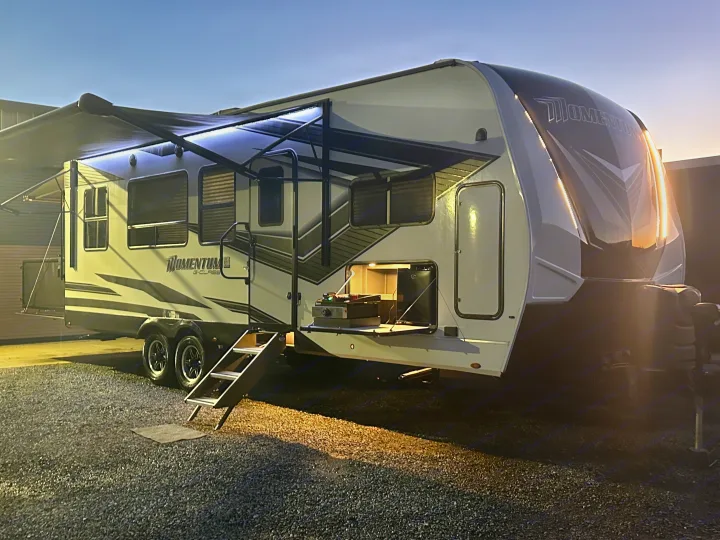 Experience comfort in this lit-up Momentum G-Class RV rental at dusk, featuring an awning, outdoor kitchen, and steps for your next adventure.. Grand Design Momentum 2022