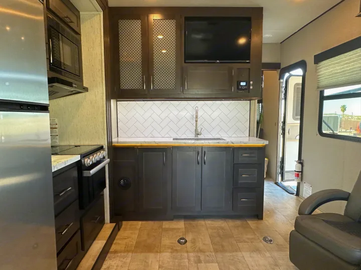 Inside a modern RV rental: sleek kitchen with dark wood cabinets, stainless steel appliances, and a stylish tiled backsplash.. Grand Design Momentum 2022