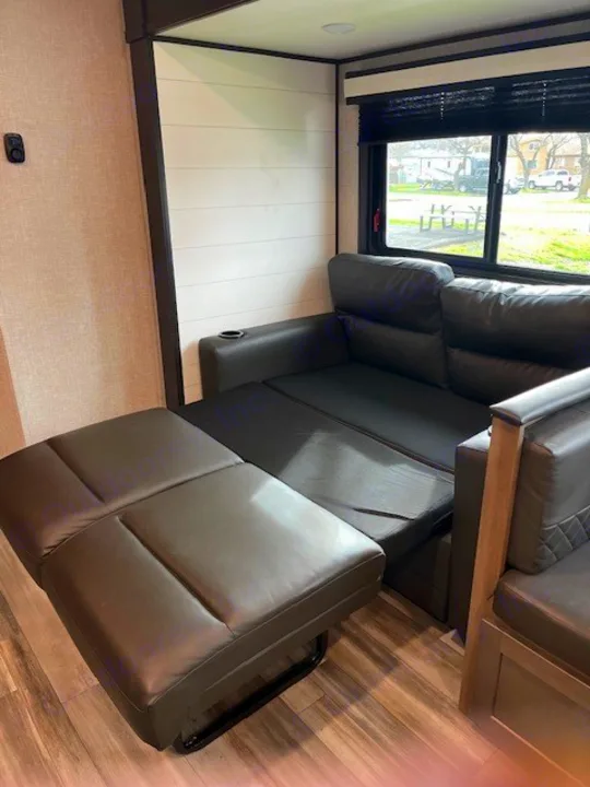 Relaxing RV rental living area with a dark, reclining sofa/bed and a view of the campsite through the window.. Jayco Jay Flight 2024