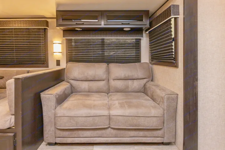 Cozy RV rental interior with a comfortable brown sofa, pleated window blinds, overhead cabinets, and wall sconce.. Jayco Jayflight 32BHDS.G 2022